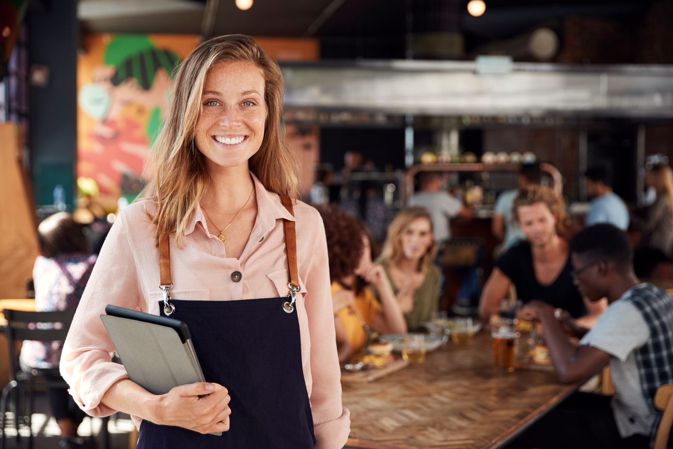 Jeune femme souriante portant un tablier dans un restaurant animé.